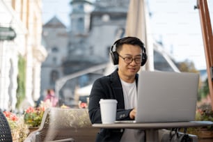 Hombre de negocios asiático con auriculares escribiendo en una computadora portátil durante el trabajo en una cafetería. Concepto de trabajo remoto y freelance. Hombre exitoso adulto enfocado con traje y gafas sentado a la mesa con café