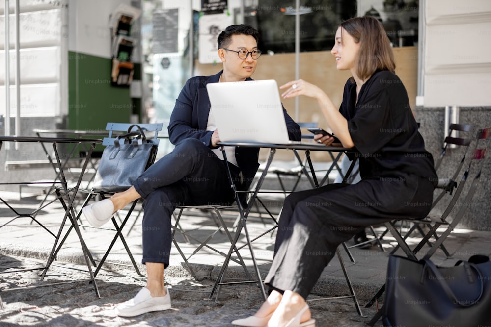 Multiracial business people watching something on laptop during work at outdoor cafe. Concept of remote and freelance work. Idea of teamwork and business cooperation. Caucasian woman with smartphone