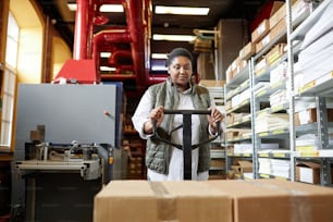 Front view portrait of female worker using pallet jack while moving boxes at printing factory warehouse
