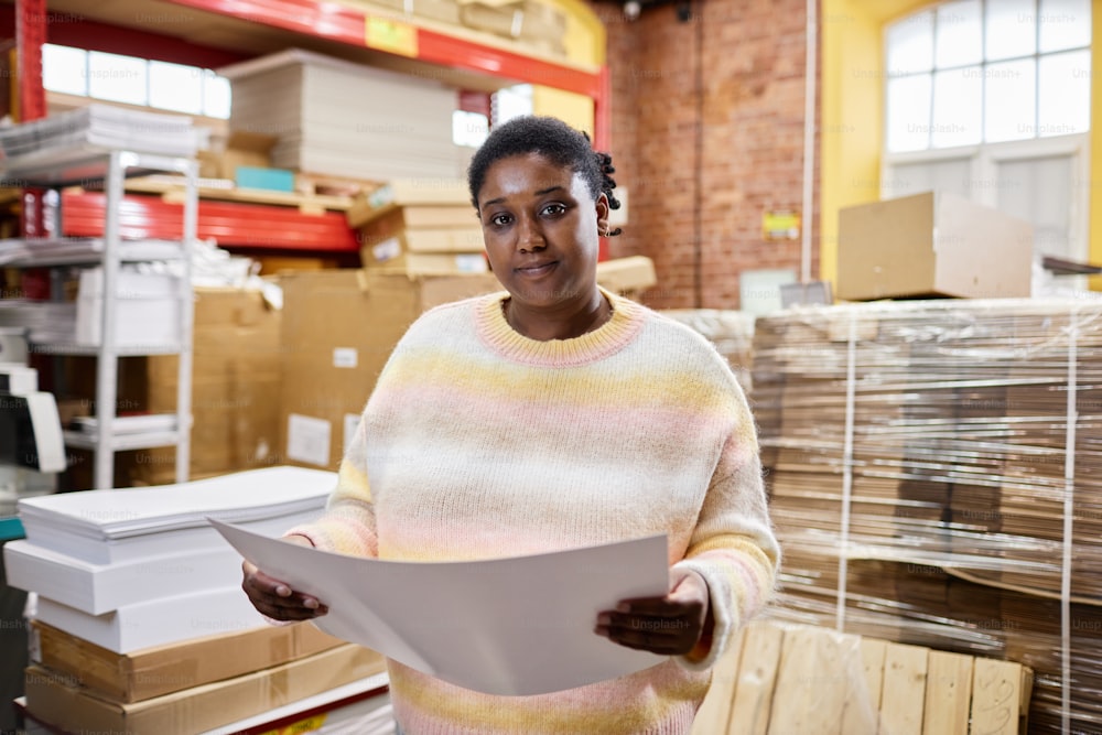 Porträt einer lächelnden schwarzen Frau, die Drucke in der Druckerei hält und in die Kamera schaut