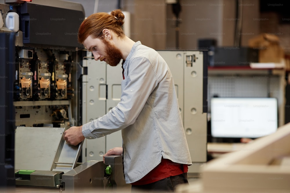Vue latérale portrait d’un jeune homme barbu travaillant dans une imprimerie et installant des machines, espace de copie
