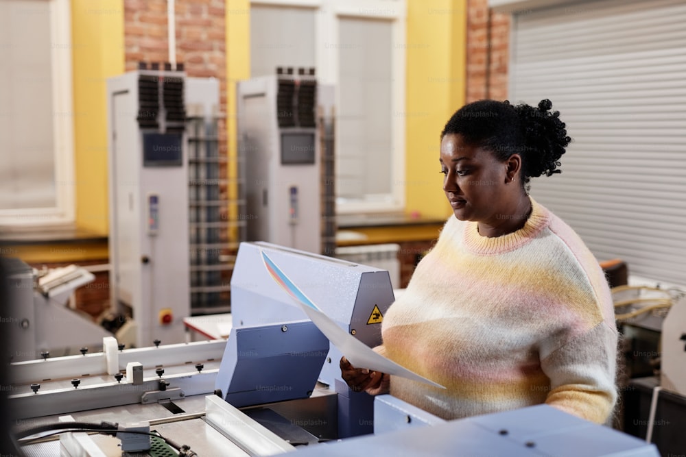 Side view portrait of young black woman operating printing machine and holding color test sheet, copy space