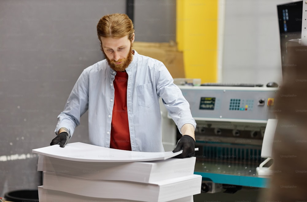 Portrait of young man working in print shop and holding stack of paper, copy space