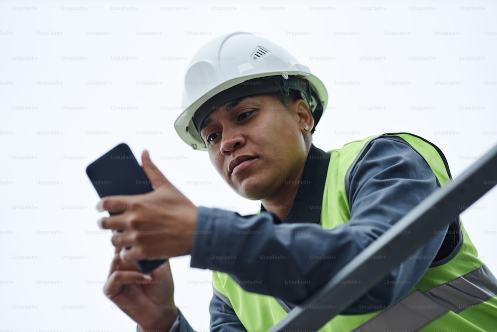 Ritratto a basso angolo di una lavoratrice che indossa l'elmetto e usa lo smartphone contro il cielo