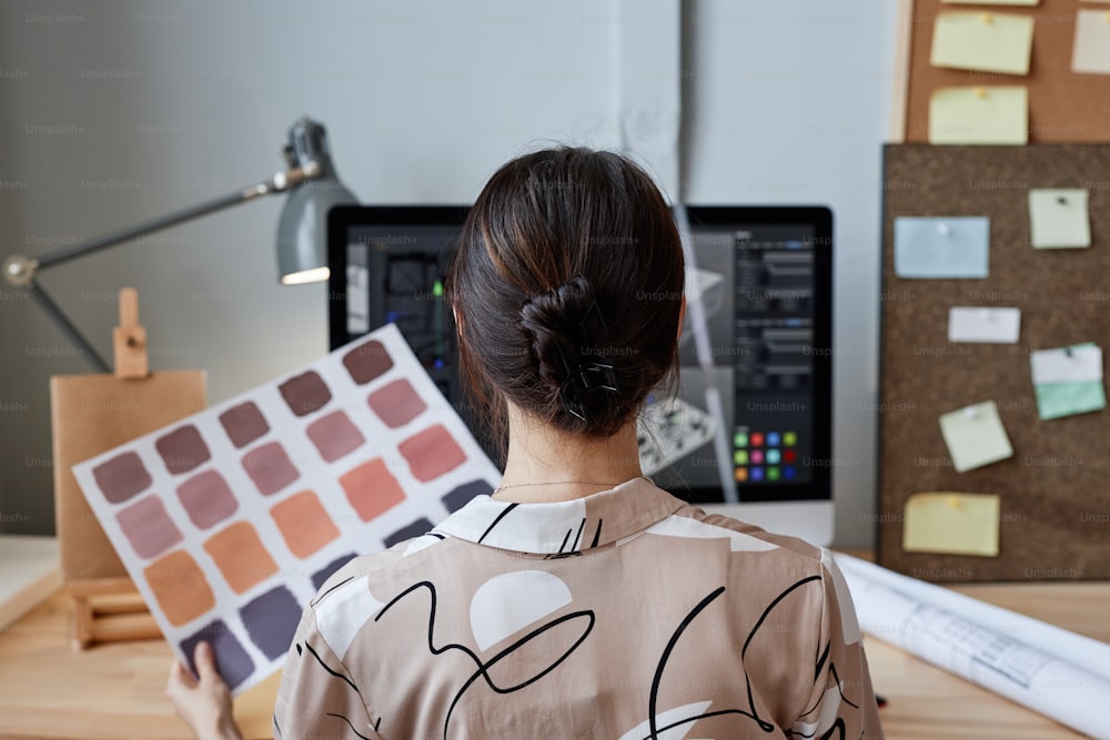 Vista traseira da designer feminina segurando amostras de cores enquanto trabalha com o computador no local de trabalho