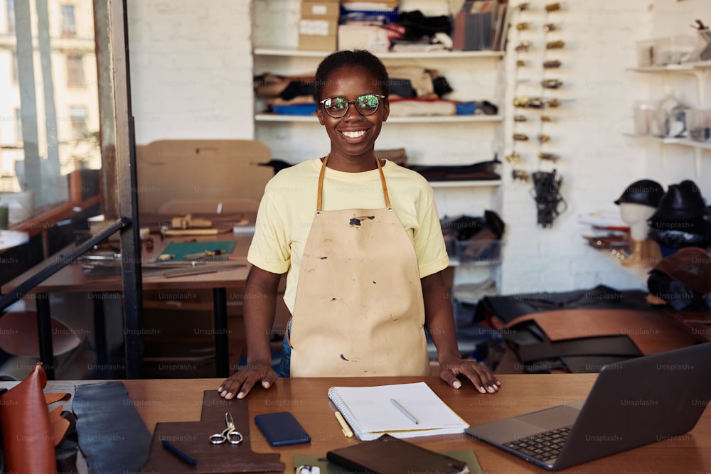 Portrait of female small business owner smiling at camera while posing in leatherworking workshop, copy space