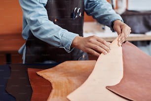 Primer plano de un joven artesano irreconocible inspeccionando cuero genuino en el taller y creando piezas hechas a mano, espacio de copia
