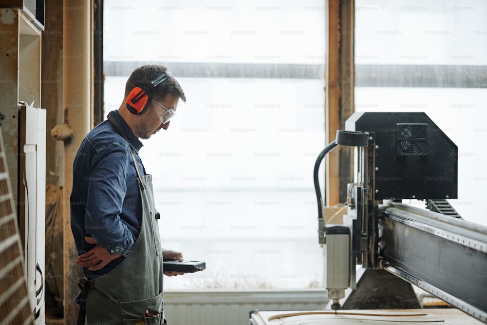 Retrato da vista lateral do carpinteiro masculino que opera a máquina de corte CNC na oficina de produção automatizada