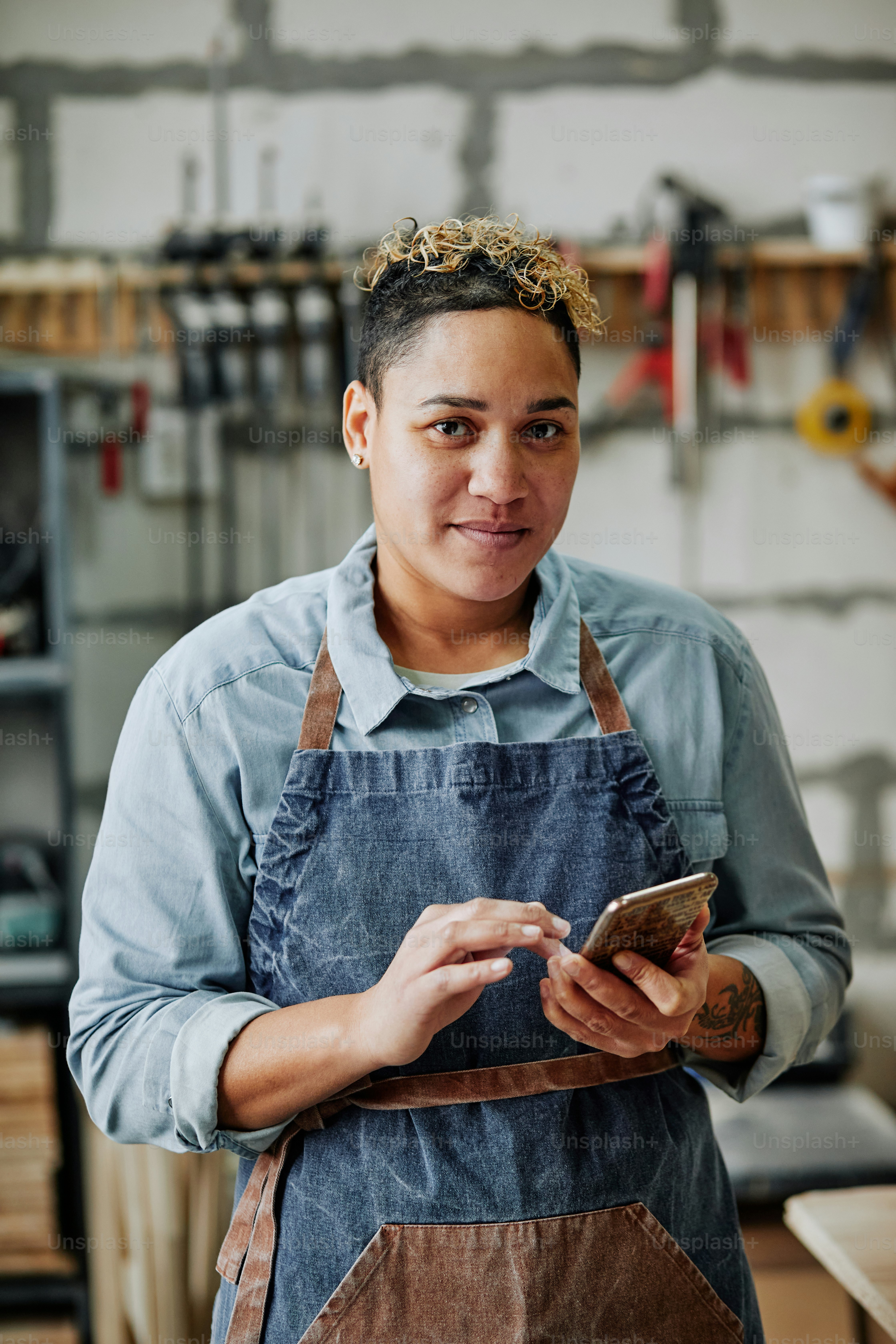 Retrato vertical da cintura para cima da artesã feminina usando o smartphone e sorrindo para a câmera enquanto está na oficina com ferramentas no fundo