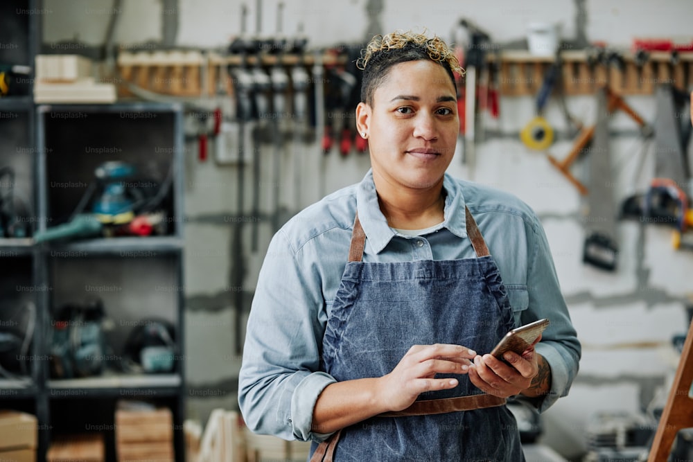 Waist up portrait of successful female artisan holding smartphone and smiling at camera while standing in workshop with tools in background, copy space