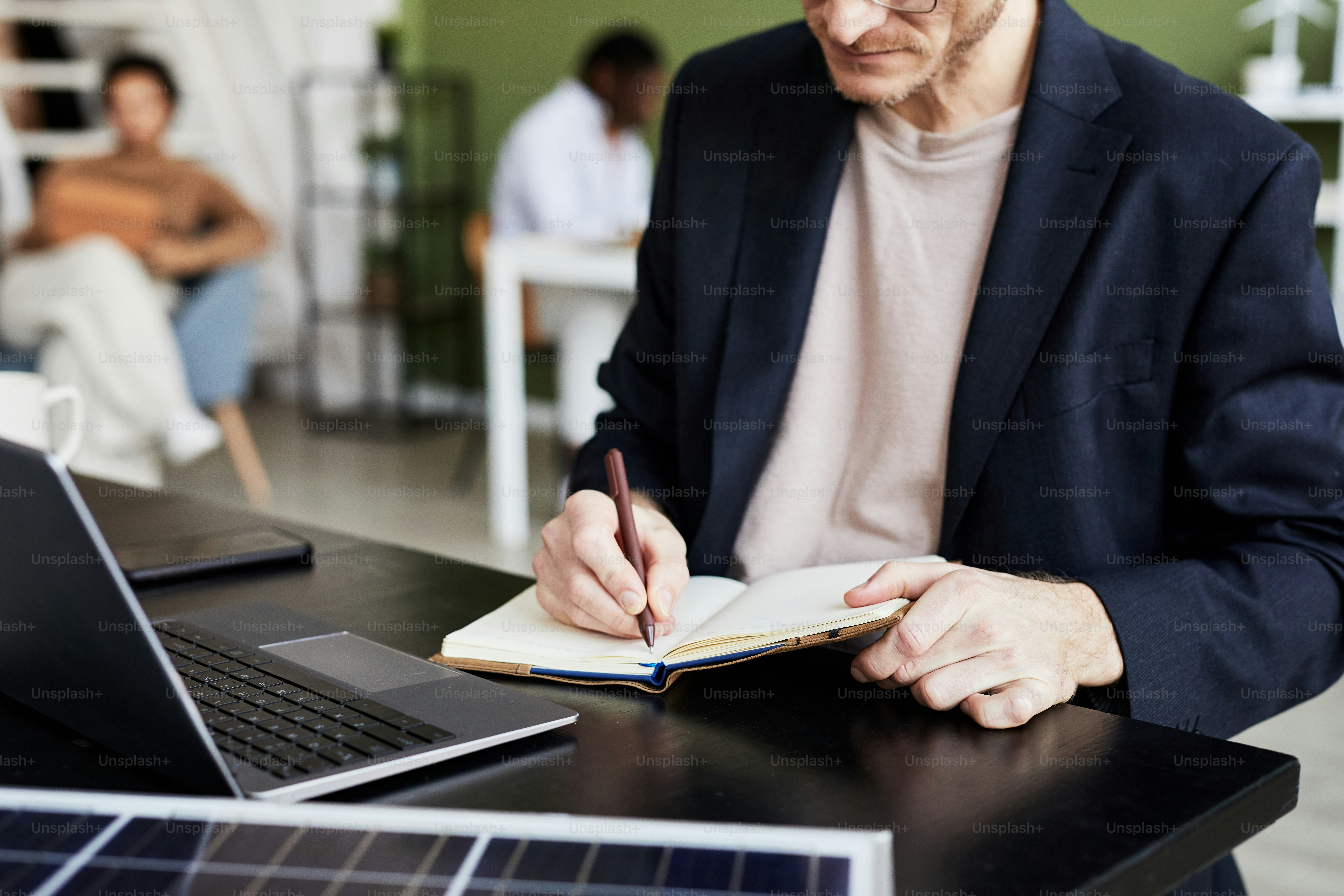 Close-up of businessman writing plans in his notebook while working at table on laptop
