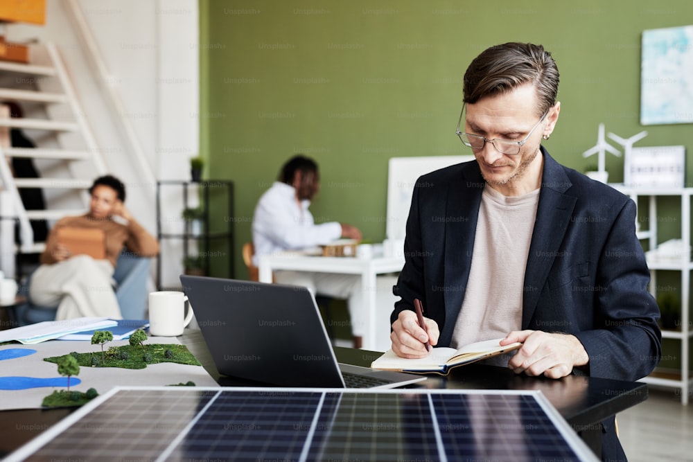 Young businessman making notes in notebook while working at table with laptop and solar panel