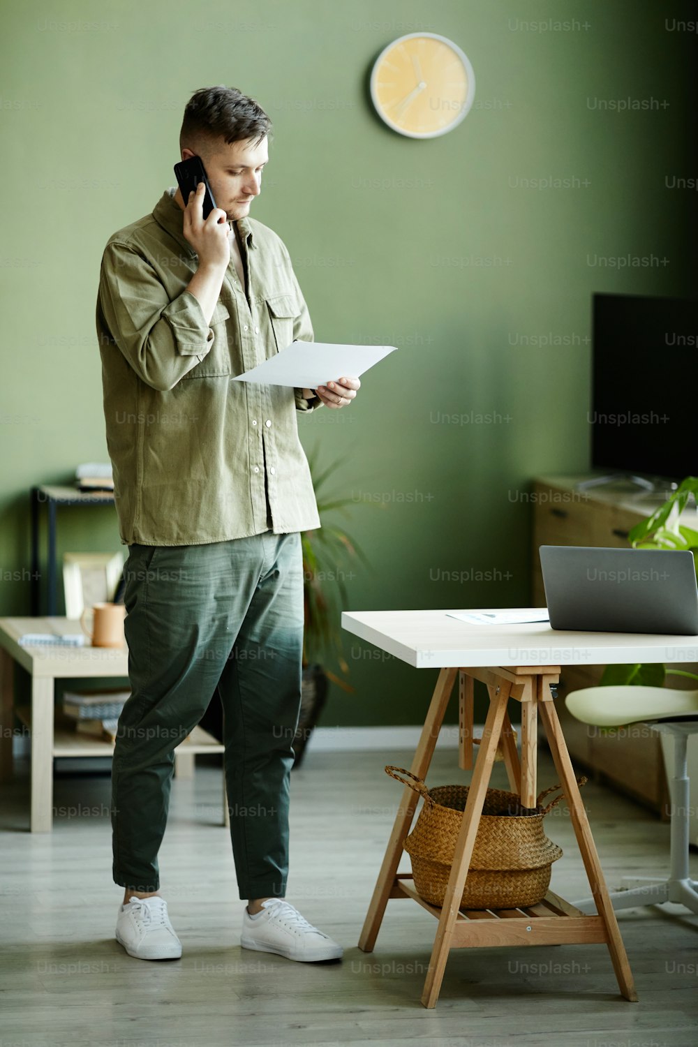 Young man discussing business document during conversation on mobile phone while standing atin the room