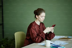 Mujer joven sentada a la mesa con documentos y escribiendo mensaje en su teléfono móvil, ella trabajando en casa