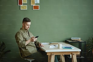 Junger Mann, der zu Hause an seinem Arbeitsplatz sitzt und eine Nachricht auf dem Handy tippt, er arbeitet online