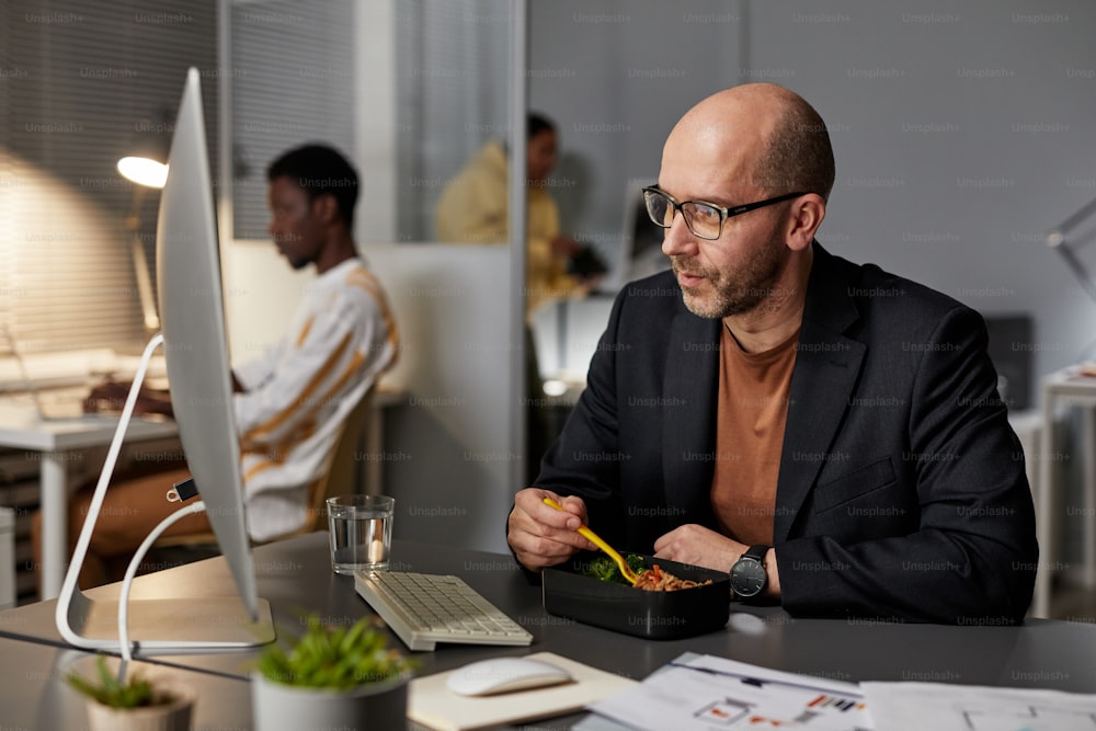 Portrait of mature businessman eating takeout dinner while working late at night in office, copy space