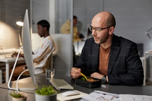 Portrait of mature businessman eating takeout dinner while working late at night in office, copy space