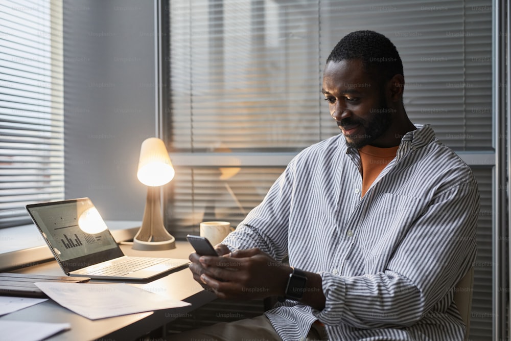 Porträt eines lächelnden schwarzen Unternehmers, der das Smartphone benutzt, während er spät im Home-Office-Arbeitsplatz arbeitet, der von einer Lampe beleuchtet wird, Kopierraum