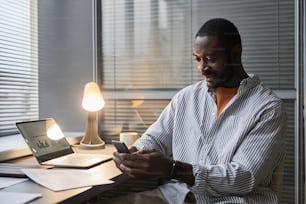 Retrato de un empresario negro sonriente que usa un teléfono inteligente mientras trabaja hasta tarde en el lugar de trabajo de la oficina en casa iluminado por una lámpara, espacio de copia