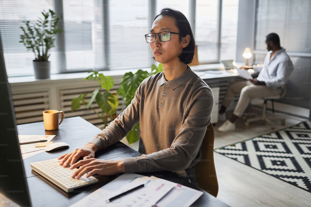 Portrait of Asian software developer using computer while working late in modern office lit by lamps