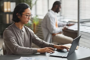 Retrato de vista lateral de un hombre asiático en ropa casual usando una computadora portátil mientras trabaja en el cubículo de la oficina, espacio de copia