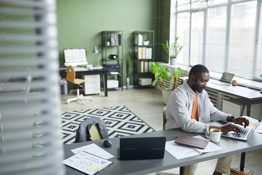 Hochwinkel-Porträt eines erwachsenen schwarzen Mannes, der im Home-Office in modernem Interieur arbeitet und einen Laptop und einen Kopierraum verwendet