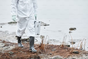 Low section shot of unrecognizable person wearing hazmat suit walking by water at ecological disaster site, copy space
