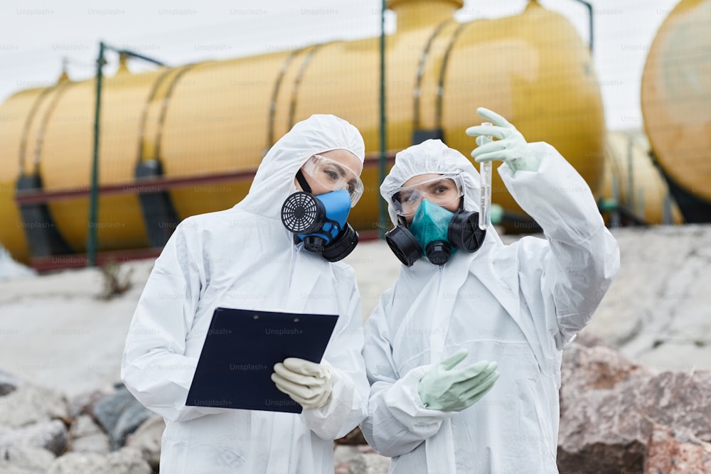Waist up portrait of two female scientists wearing hazmat suits collecting samples outdoors, toxic waste and pollution concept
