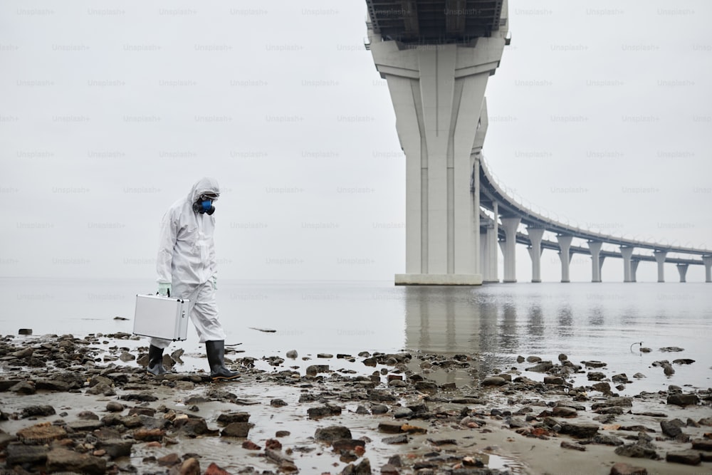 Graphic wide angle portrait of man wearing hazmat suit collecting probes by water, toxic waste and pollution concept, copy space
