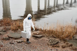 Retrato de corpo inteiro do trabalhador vestindo o traje de hazmat andando pela água ao ar livre carregando o caso de amostras, conceito de resíduos industriais, espaço de cópia