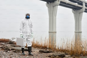 Retrato do trabalhador vestindo o traje de hazmat andando pela água ao ar livre e carregando o caso de amostras, conceito de resíduos industriais, espaço de cópia