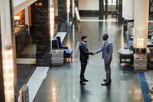 Wide angle view at two business partners shaking hands while standing in luxurious hotel lobby, copy space