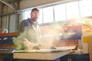 Lächelnder bärtiger Fabrikarbeiter mit Schutzbrille, der an der Werkbank steht und mit Spanplatten in staubiger Werkstatt arbeitet