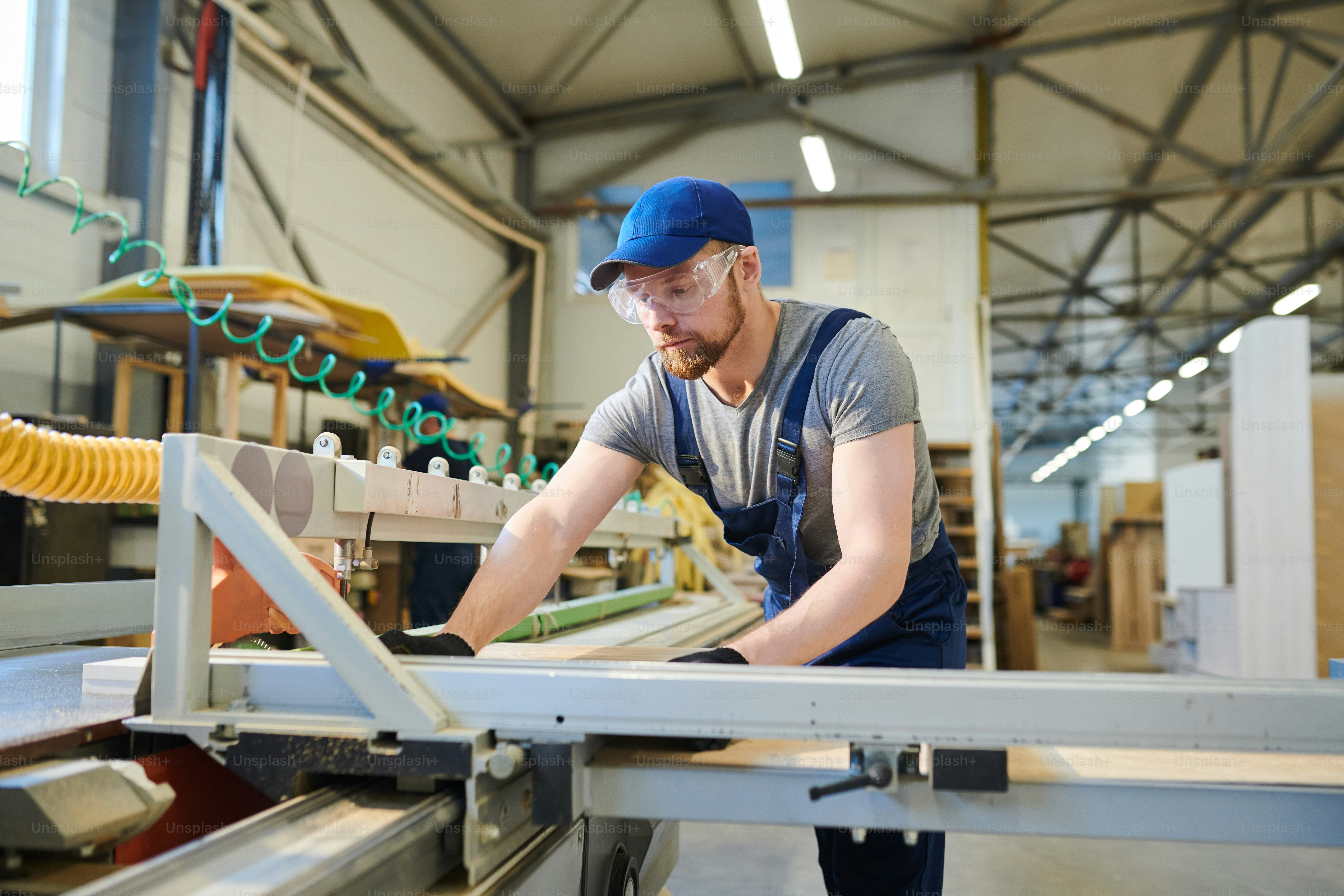 Serious concentrated bearded engineer in blue cap and safety goggles adjusting wooden pieces while  joining furniture details on modern machine