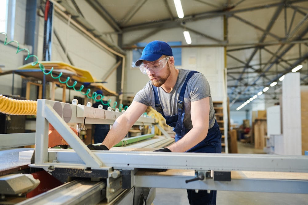 Engenheiro barbudo concentrado sério em boné azul e óculos de segurança ajustando peças de madeira enquanto une detalhes de móveis na máquina moderna