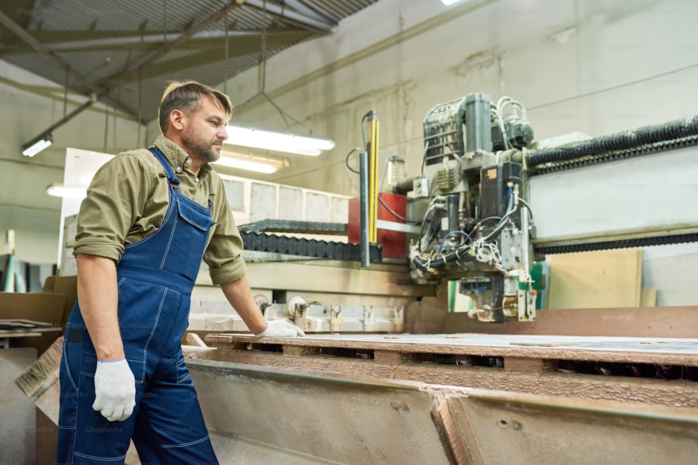 Retrato de vista lateral de un trabajador maduro de pie junto a la máquina de corte de madera en una fábrica de muebles modernos, espacio de copia