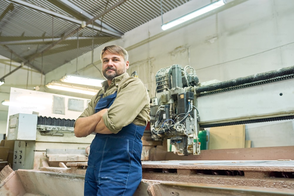 Retrato do trabalhador maduro sorridente posando com os braços cruzados e olhando para a câmera pela máquina de corte de madeira na fábrica de móveis moderna, espaço de cópia