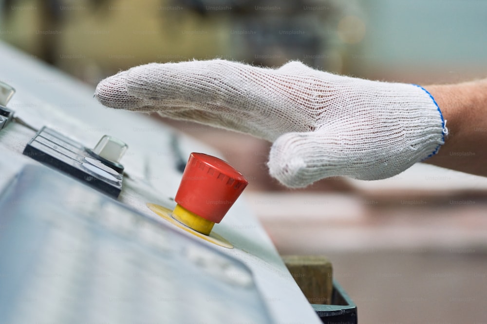 Vue latérale rapprochée de la main masculine dans un gant de travail appuyant sur le gros bouton rouge STOP en cas d’urgence