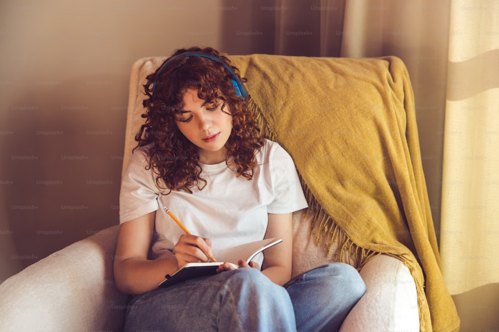 Diary. Curly-haired young girl in headphones sitting in the armchair and making notes in a diary