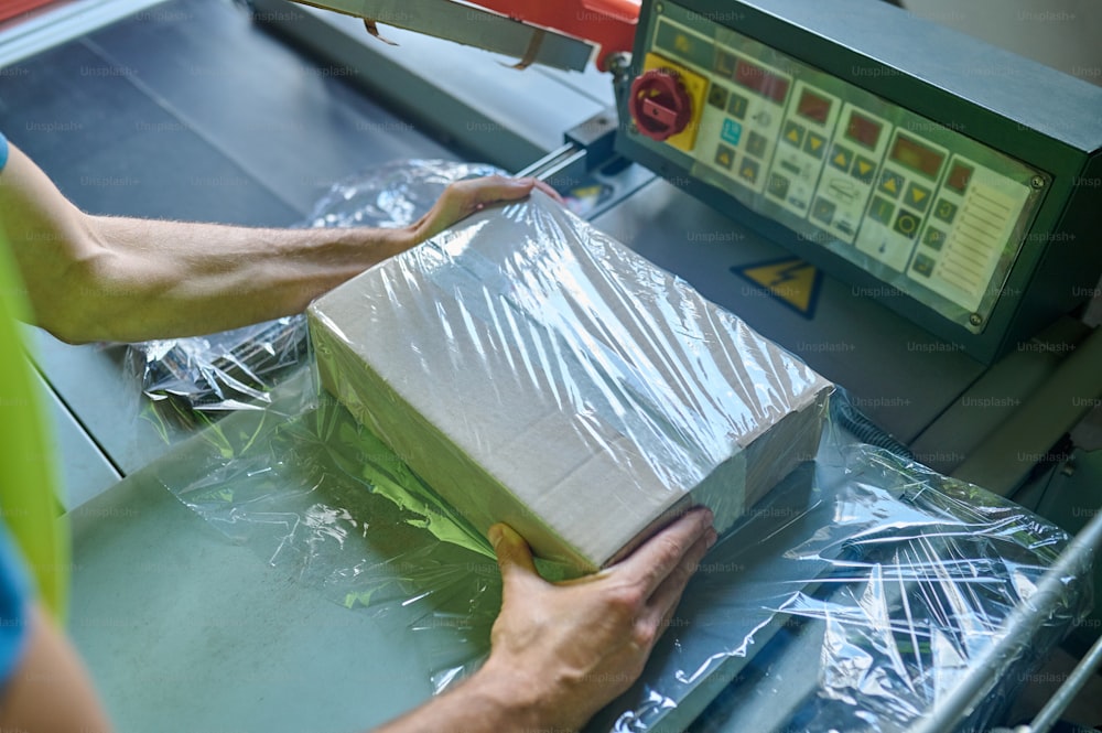Photo recadrée d’un employé d’entrepôt emballant la boîte en carton avec la machine d’emballage à film étirable