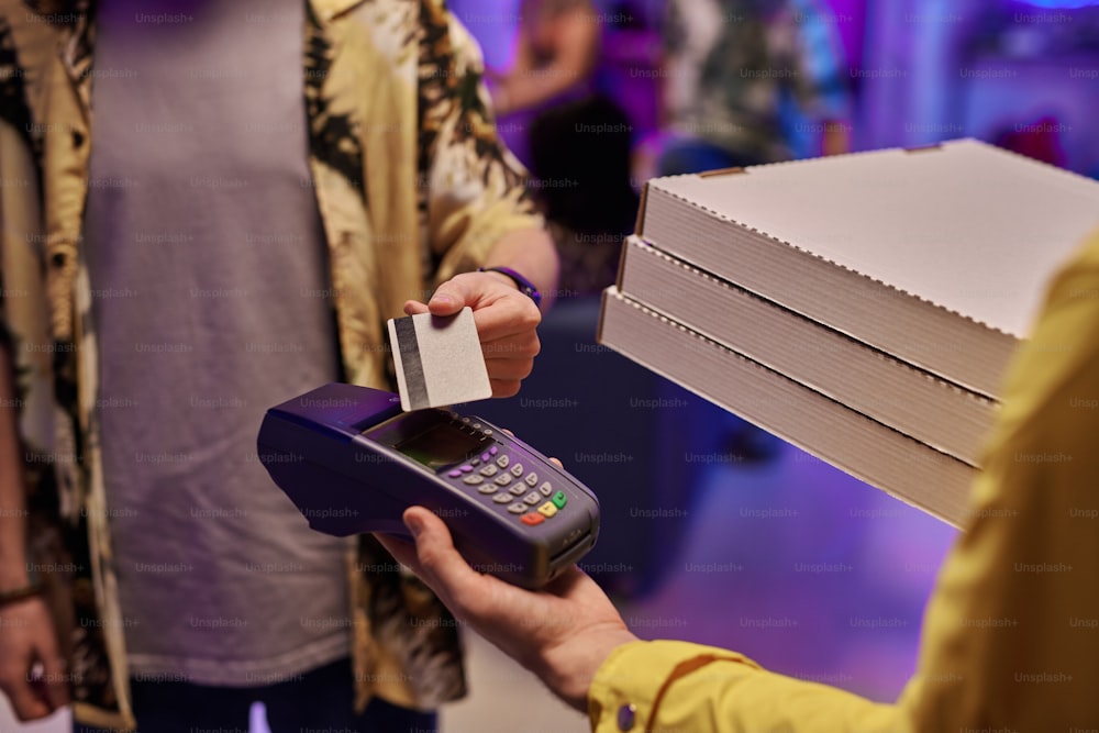 Jeune homme en tenue décontractée payant par carte de crédit la pizza commandée emballée dans des boîtes en carton carrées tenues par un livreur en uniforme