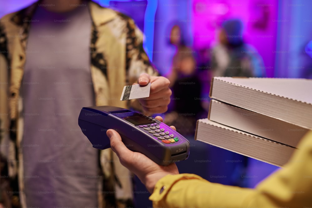 Hand des Kuriers, der Kisten mit Pizza und Zahlungsterminal hält, während der Typ die Kreditkarte benutzt, um das bestellte Essen zu bezahlen, um es zu Hause zu essen Party