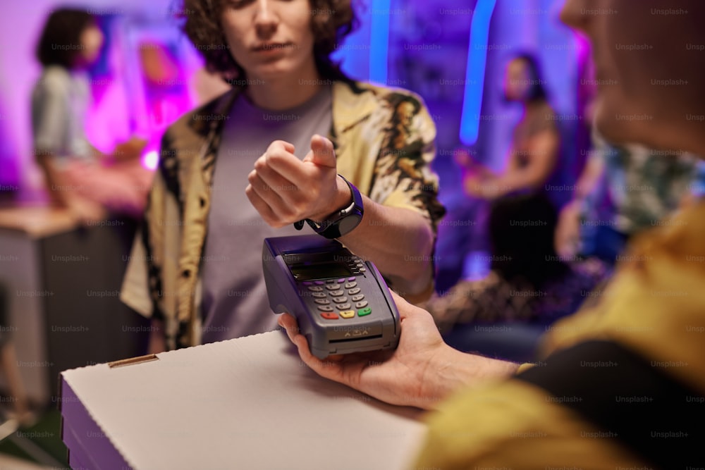 Main du gars avec smartwatch sur le terminal de paiement et les boîtes avec pizza tenues par le livreur contre un groupe d’amis profitant de la fête à la maison