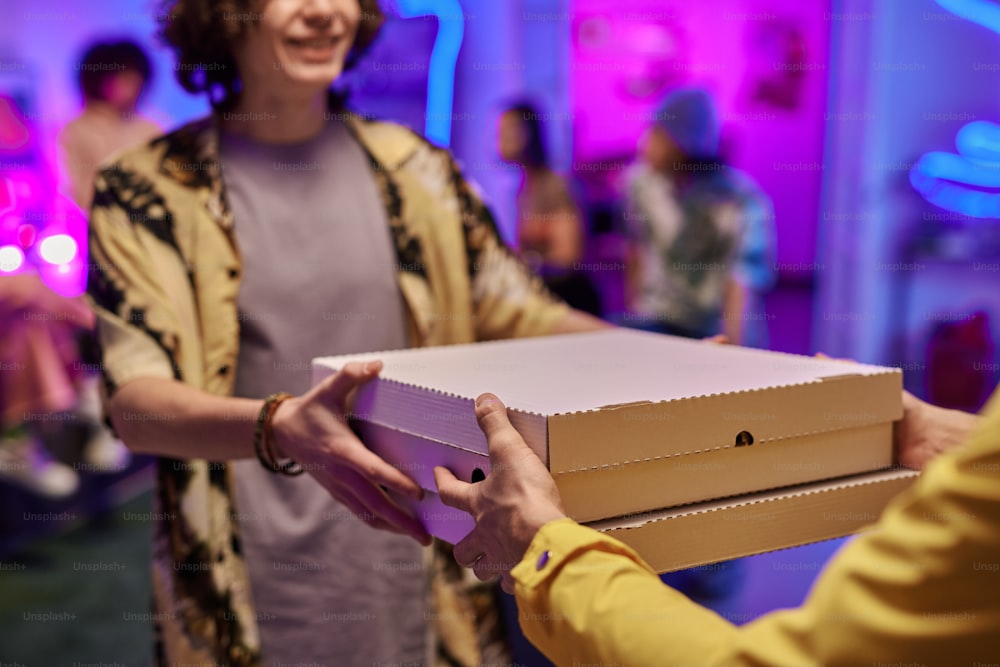 Gros plan d’un jeune homme souriant prenant des boîtes avec des pizzas passées par un livreur en uniforme contre un groupe d’amis faisant la fête