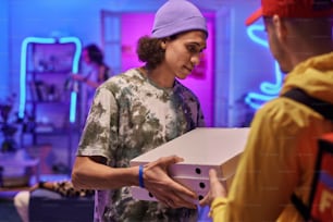 Joven en ropa casual sosteniendo una pila de cajas de cartón cuadradas con pizza mientras está parado frente al mensajero durante la fiesta en casa