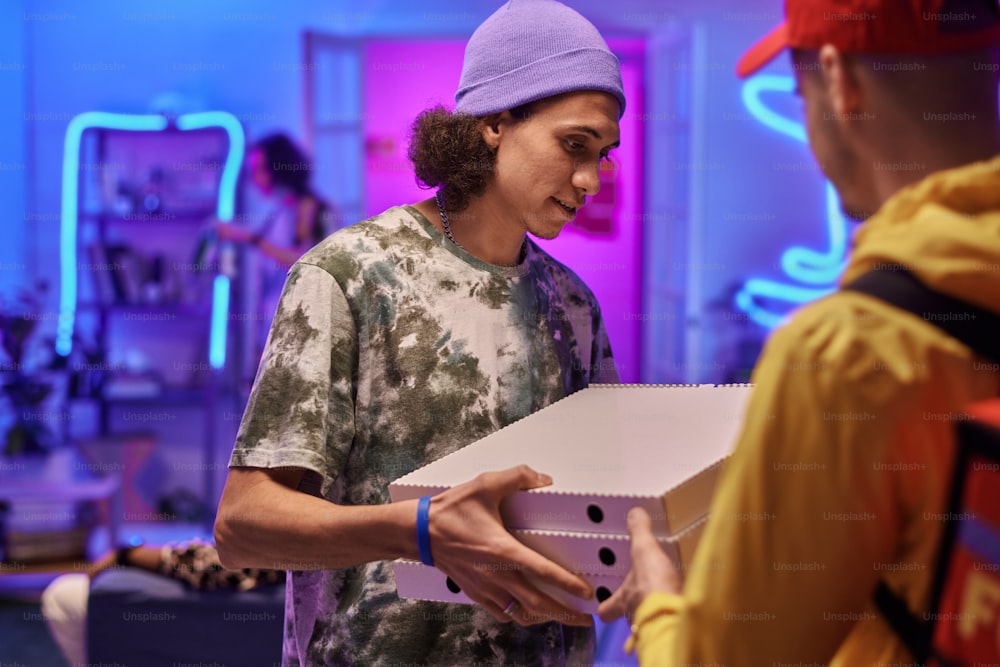 Young man in casualwear holding stack of square cardboard boxes with pizza while standing in front of courier during home party