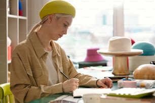 Side view of young creative hat designer drawing sketches of new models for seasonal fashion collection in notebook by her workplace