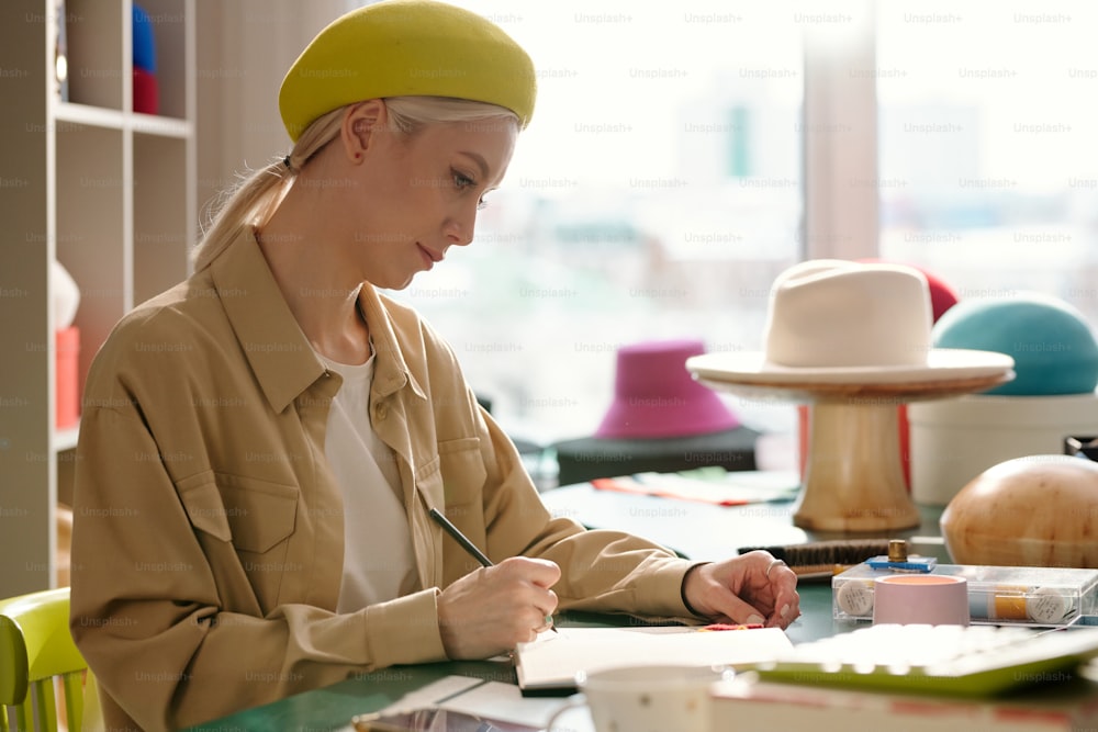 Vista lateral de una joven diseñadora creativa de sombreros dibujando bocetos de nuevos modelos para la colección de moda de temporada en cuaderno por su lugar de trabajo