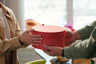 Close-up of young female vendor giving red round box with packed handmade product to client of her craft shop during work