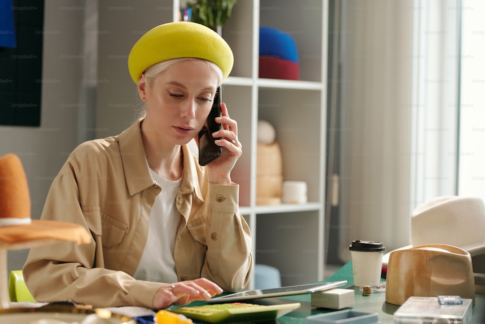 Jeune femme entrepreneur en tenue décontractée élégante parlant sur un téléphone portable et appuyant sur les boutons de la calculatrice tout en travaillant en studio